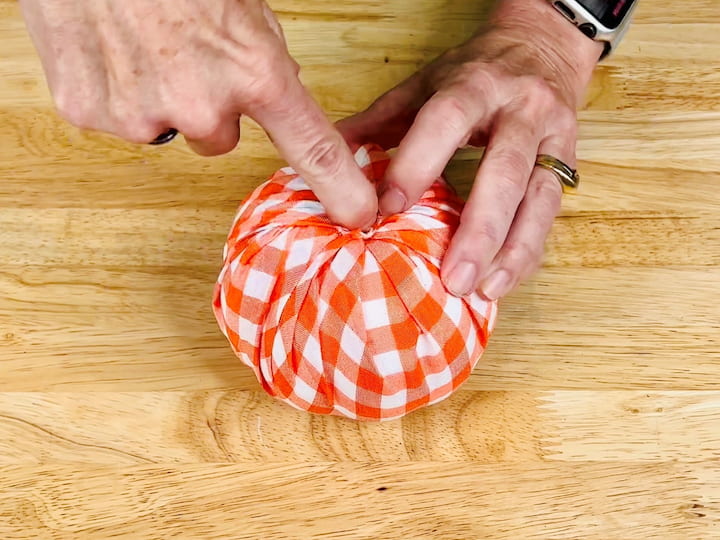 Place the styrofoam pumpkin in the center of the fabric circle. Begin pulling up the fabric around the pumpkin, tucking it into the center. Continue until the entire pumpkin is covered with fabric, creating folds and gathers.