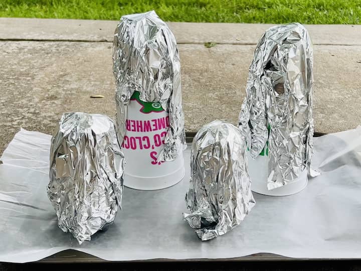 Cover your work surface with wax paper.  Place cups and water bottles on the wax paper. Create tin foil balls and place them on top of each cup or bottle. Wrap tin foil around the entire structure.