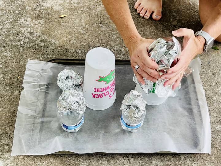Cover your work surface with wax paper.  Place cups and water bottles on the wax paper. Create tin foil balls and place them on top of each cup or bottle. Wrap tin foil around the entire structure.