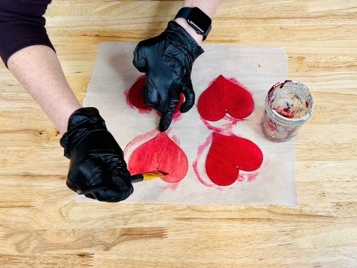 I'll be using Folk Art Ultra Dye and I covered my hands with gloves to protect them from being stained or dyed. I poured a little bit of the dye into a container and then I used a paintbrush to paint it onto the wooden hearts. I went ahead and gave the hearts two coats so they'd be a nice vibrant color and then let them dry completely. 