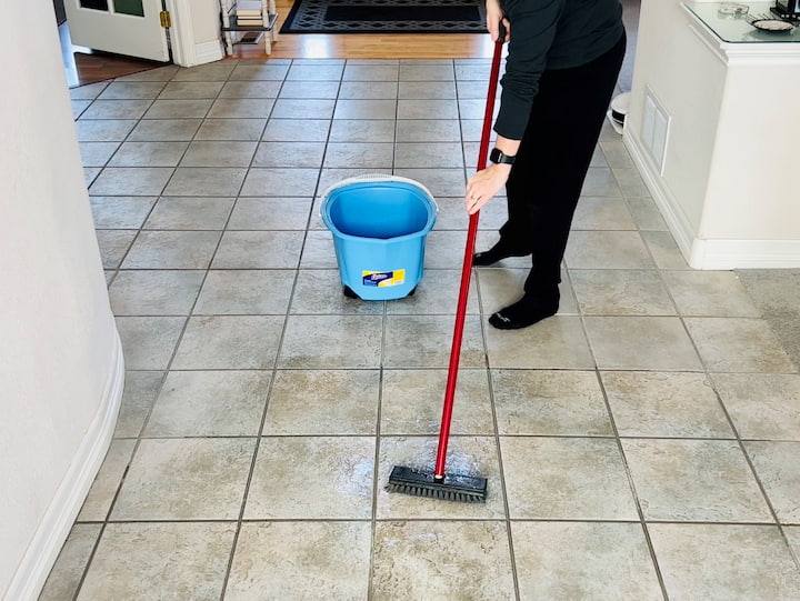 What we're going to go ahead and do now is take our bucket out and get a deck brush and you're going to dip the deck brush into the vinegar water and then you're going to put it onto your tile and you will scrub it back and forth. This is going to clean your tile scrub it into the pores and clean your grout all at the same time.