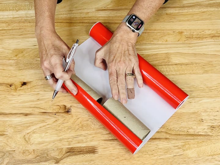 Measure 2 toilet paper rolls with red vinyl, cut the vinyl, stick it to the tubes, and glue the red tubes on the sides.